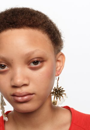 Joven con cabello corto y rizado, lleva una camiseta roja y un gran pendiente dorado en forma de sol con cuentas blancas colgantes en la oreja izquierda.