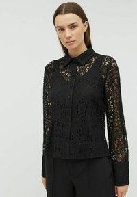 Sort blonde bluse med knaplukning foran, spids krave og lange ærmer, der har blomstermønstre og en tekstureret overflade.