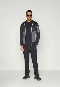 Must küllane treeningjakk hallide aktsentidega, esime silmavärava ja taskutega, koos mustade pükste ja kingadega. Mudel kannab päikeseprille.