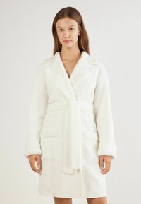 Vit badrock med en texturerad tyg, mjuk fuskpälskrage och manschetter, bälte i midjan och två framläggande fickor.