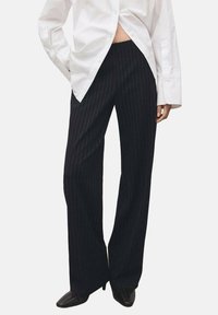 Zwarte pantalon met krijtstreep en een losse pasvorm, gemaakt van een gladde stof. De tailleband zit boven de heupen en wordt gecombineerd met een lang wit overhemd.