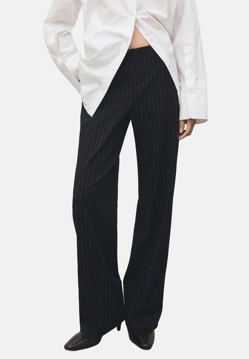 Zwarte pantalon met krijtstreep en een losse pasvorm, gemaakt van een gladde stof. De tailleband zit boven de heupen en wordt gecombineerd met een lang wit overhemd.