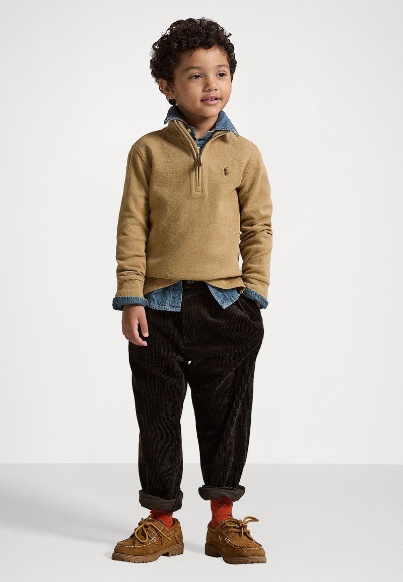 Beige stripete genser med glidelås, denimkrage. Brune kordfløyelsbukser med oppbrett i ermene. Brune sko med dekorative snørebånd, røde sokker med mønster.