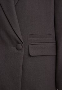 Blazer gris oscuro con un tejido texturizado, cierre de un solo botón y un bolsillo horizontal en el lado izquierdo.