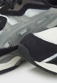 Schwarze und graue Sportschuhe mit Netzpaneelen, Wildlederakzenten und einer strukturierten Sohle. Das Design umfasst eine dicke, gepolsterte Zwischensohle.