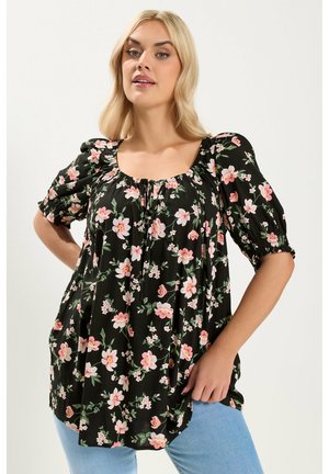 Vaaleahiuksinen nainen pukeutuneena mustaan blousuun, jossa on vaaleanpunainen kukkakuvio ja puffihihat, yhdistettynä vaaleansinisiin farkkuihin, poseeraa yksinkertaisen taustan edessä.