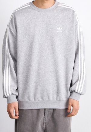 Mand iført grå Adidas sweatshirt med hvide striber på ærmerne og lille logo på brystet, kombineret med grå manchesterbukser.