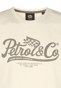 Cremefarvet bomulds t-shirt med mørkegrå tekst: "Petrol & Co" med et ternet flag-design og "Denim Driven Workwear" nedenunder.