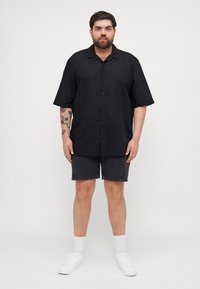 Svart kortermet skjorte med knappelukking, kombinert med mørke shorts. Modellen har på seg hvite joggesko og sokker, med synlige tatoveringer på armene.