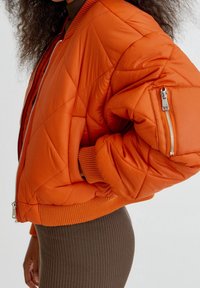 Veste bomber orange matelassée avec fermeture éclair, poignets côtelés et poche latérale zippée, présentant une finition lisse et brillante.