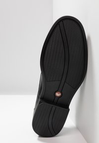 Musta kumisaapas, jossa on teksturoitu pinta ja kaareva muoto, keskellä logo-yksityiskohta. Selvä pohjakuviot näkyvissä.