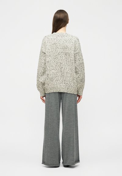 Person mit langen braunen Haaren trägt einen gesprenkelten hellgrauen Pullover und weit geschnittene graue Hosen mit weitem Bein, steht mit dem Rücken zur Kamera.