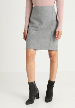 Kobieta w szarej spódnicy do kolan, jasnoróżowej bluzce z długim rękawem oraz czarnych zamszowych botkach na słupku na jednolitym tle.