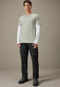 Grå kortærmet t-shirt over en hvid langærmet skjorte, parret med sorte cargo-bukser og sorte vandresko, stående foran en neutral baggrund.