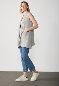 Light grey sleeveless vest with open front, side pockets, paired with light-wash denim jeans and white sneakers with brown accents.