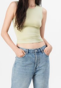 Kobieta w jasnooliwkowej bluzce bez rękawów i niebieskich dżinsach z wysokim stanem, z dłońmi w kieszeniach, na białym tle.