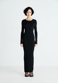 Abito lungo nero a coste con maniche lunghe, scollo rotondo e silhouette aderente; tessuto leggero; indossato con sandali neri chunky.