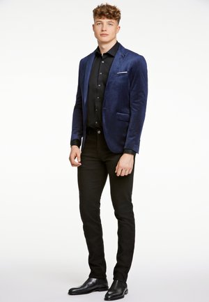 Marineblauwe fluwelen blazer met geknipte revers, enkele zak; zwart shirt en slim zwarte broek; zwarte enkellaarsjes maken de outfit compleet.