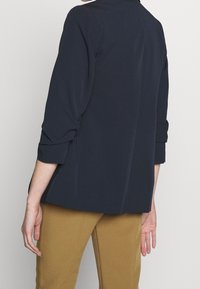 Námořnická modrá, lehká textilní bunda s tříčtvrtečními rukávy, s volným střihem a detailem švu na zadní části. V kombinaci s béžovými kalhotami.