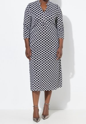 Vrouw draagt een zwart-wit midi-jurk met een geometrisch patroon, driekwart mouwen en zilveren puntige hakken met enkelbandjes.