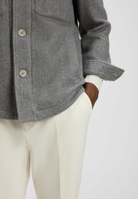 Gray herringbone jacket with two front pockets and button closure, paired with cream-colored trousers, featuring a cuffed sleeve design.