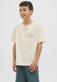 Cremefarbenes T-Shirt mit kurzen Ärmeln und lockerem Schnitt, mit einem kleinen Logo in schwarzem Text auf der linken Brustseite, kombiniert mit hellblauen Shorts.