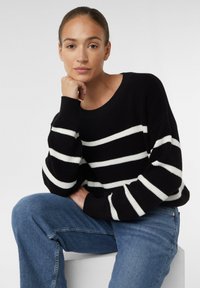 Sweter z czarno-białymi paskami, wykonany z dzianiny o żeberkowej fakturze i szerokim dekolcie, zestawiony z niebieskimi jeansami. Luźny fason, casualowy design.