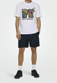 Uomo che indossa una maglietta grafica bianca con il logo MTV, pantaloni corti neri, calze bianche e scarpe da ginnastica nere, in piedi davanti a uno sfondo bianco semplice.