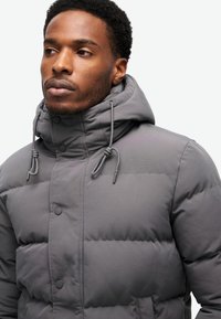 Chaqueta gris acolchada con cuello alto, capucha ajustable con cordón y botones a presión. Presenta una textura suave y costuras uniformes.