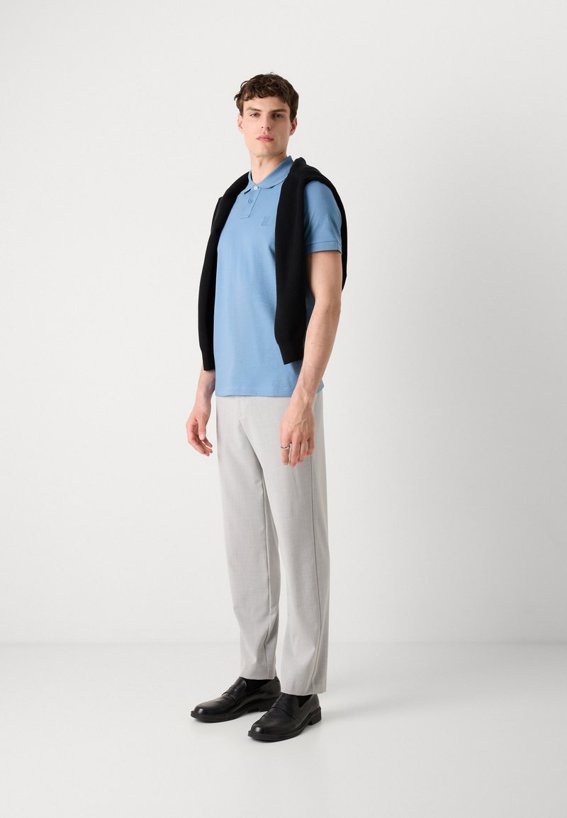 Camisa polo azul claro con un logo sutil, suéter negro caído sobre los hombros, pantalones grises claros a medida y zapatos de vestir negros.