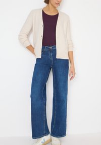 Cárdigan de punto beige con escote en V, camiseta morada debajo y vaqueros de pierna ancha azul. El modelo lleva zapatillas blancas con detalles en azul claro.