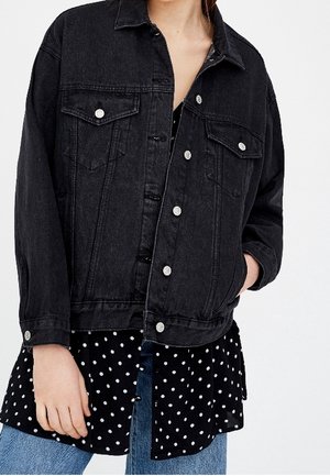 Zwarte spijkerjas met zilverkleurige knopen, gedragen over een zwarte polkadotblouse en blauwe spijkerbroek, hand rustend in het jaszak.