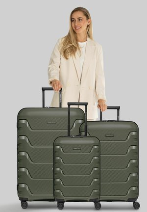 Vrouw in beige blazer staande achter drie olijfgroene harde koffers met uitgetrokken handvatten op een grijze achtergrond.