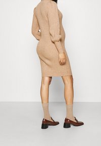 Robe moulante tricotée couleur marron clair avec texture côtelée. Présente des manches bouffantes, une coupe mi-longue, et est assortie de chaussures vernies marron et de chaussettes beiges.