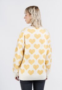 Maglione crema con motivo a cuore giallo, texture lavorata a maglia, spalle basse e polsini a costine. Presenta una vestibilità rilassata e oversize.