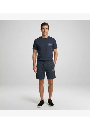 Man draagt een marineblauw T-shirt, bijpassende short met trekkoord en tekst, en zwarte sneakers, staand tegen een effen lichtgrijze achtergrond.