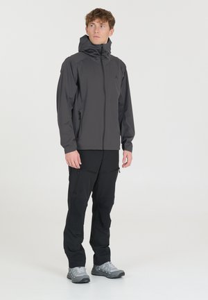 Veste grise imperméable pour homme avec capuche, fermeture éclair intégrale et poches latérales. Portée avec un pantalon noir et des chaussures de randonnée grises. Texture lisse.