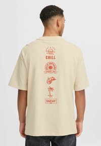 Šviesiai smėlinė trumpa rankovė marškinėliai su raudonomis grafikomis ant nugaros, įskaitant saulę, palmę ir tekstus: "CHILL," "VACAY" ir kt.