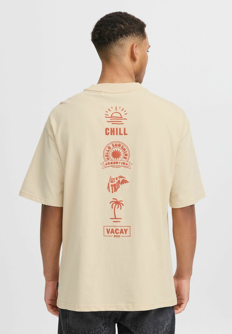 Beige tričko s krátkým rukávem, které má červené grafiky na zadní straně, včetně slunce, palmy a textu: "CHILL," "VACAY," a dalších.
