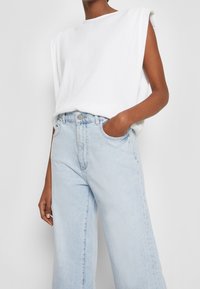Top sin mangas de canalé blanco combinado con jeans de pierna ancha de tiro alto en azul claro. Los jeans cuentan con bolsillos frontales y un cierre de botón sencillo.