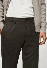 Donkergroene broek van gladde stof met een vlakke voorkant, zijzakken en een knoopsluiting, gecombineerd met een lichtgrijs vest.