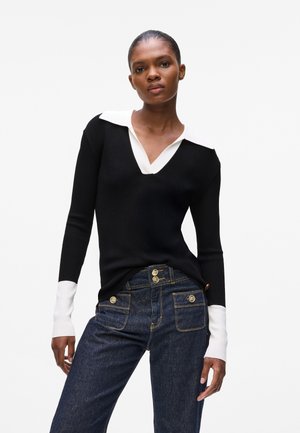 Black ribbed sweater with a white collared neckline and long sleeves; worn with dark blue denim jeans featuring gold buttons and pockets.
