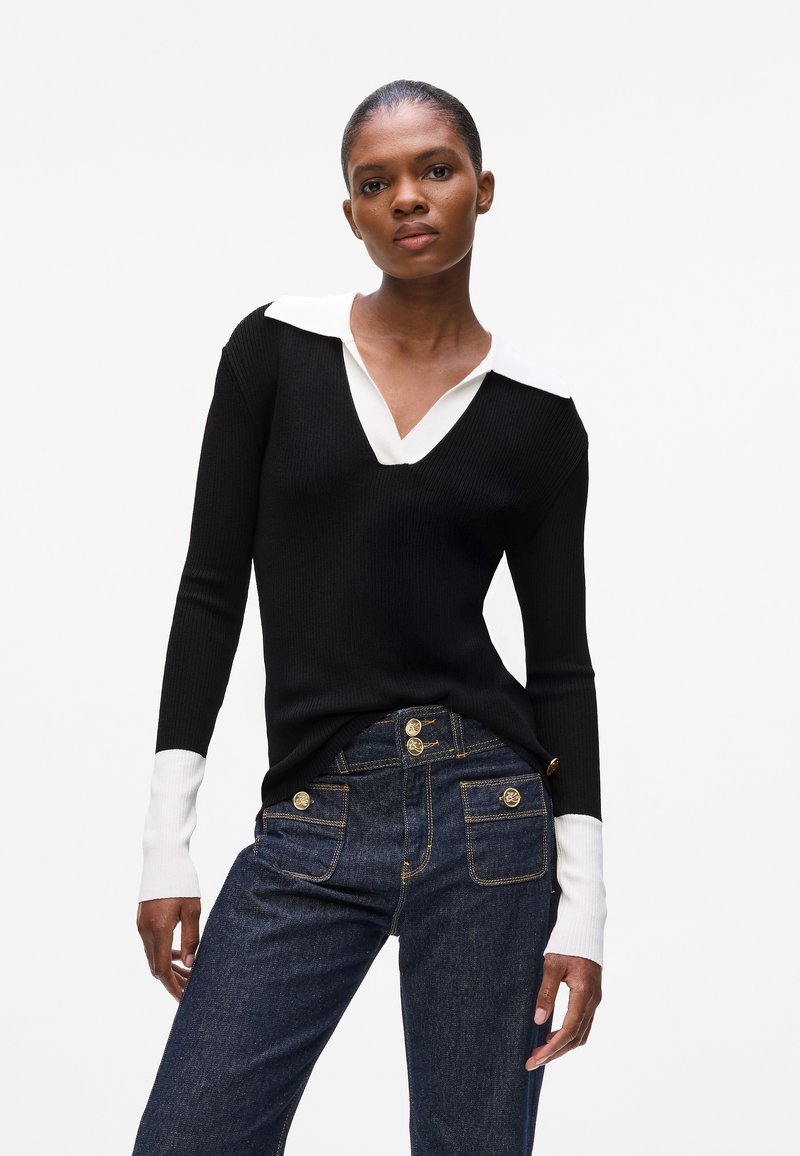 Zwarte geribbelde trui met een witte kraag en lange mouwen; gedragen met donkerblauwe jeans van denim met gouden knopen en zakken.