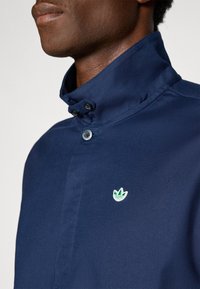 Close-up de uma pessoa a usar uma camisa azul-marinho abotoada com um colarinho alto e um pequeno logotipo bordado verde e branco no peito.