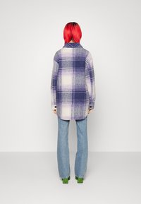 Person mit leuchtend rotem Haar, der eine lila-weiße kariert Jacke, hellblaue Jeans und grüne Schuhe trägt und mit dem Rücken zur Kamera steht.