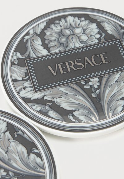 Δύο διακοσμητικά πιάτα με περίτεχνα γκρι και λευκά λουλουδένια σχέδια· ένα πιάτο έχει ένα ορθογώνιο ετικέτα με το κείμενο "VERSACE".