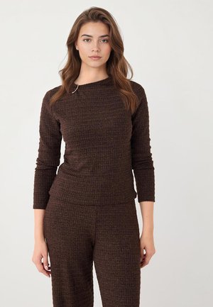 Vrouw met lang golvend haar, gekleed in een bijpassende donkerbruine, getextureerde gebreide top en broek, staand tegen een effen lichte achtergrond.