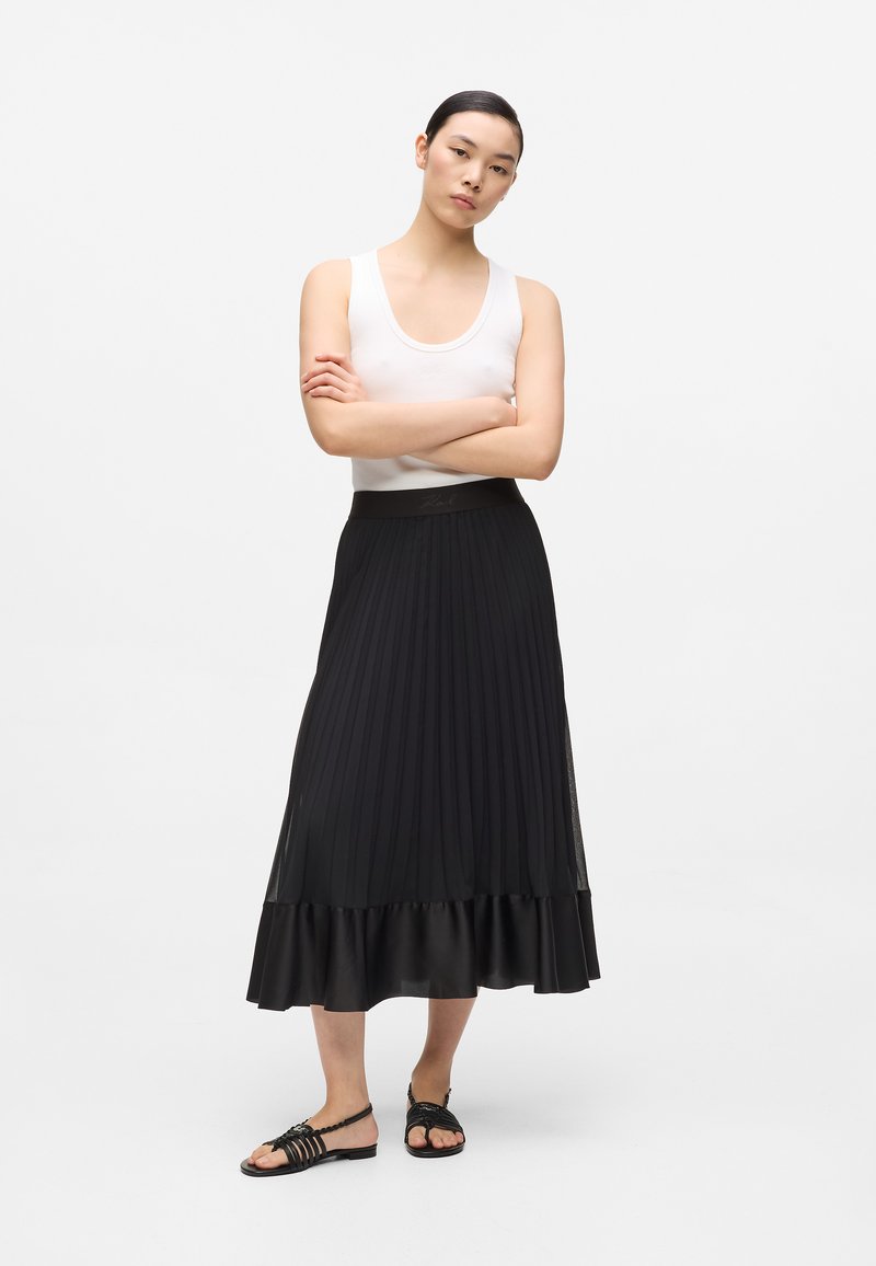 Jupe plissée noire avec un ourlet en satin, associée à un débardeur blanc ajusté. Le modèle porte des sandales noires à lanières.