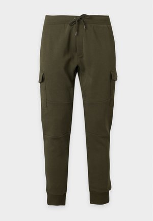 Erwachsener Mann, der ein braunes Langarmhemd, olivgrüne Cargo-Hose und weiße Sneakers trägt und vor einem schlichten weißen Hintergrund steht.