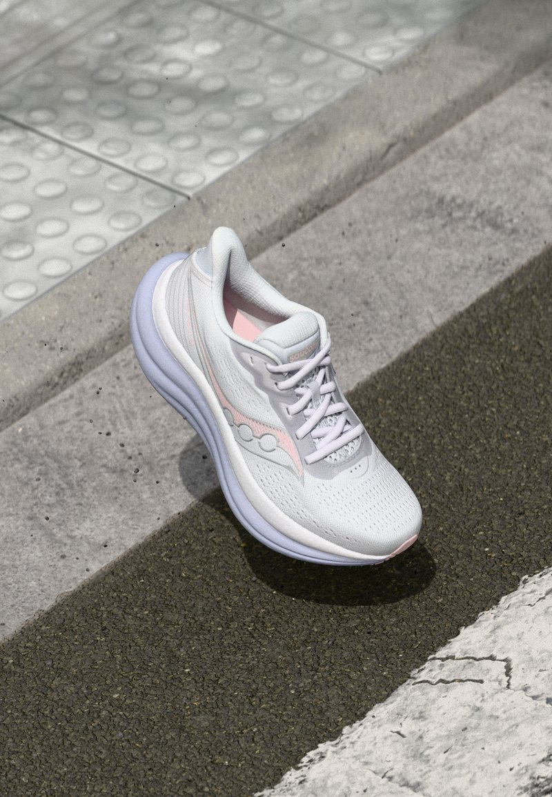 Chaussure de sport blanche avec une tige en mesh respirant, des accents roses et une semelle rembourrée. Présente un design à lacets et un logo sur le côté.